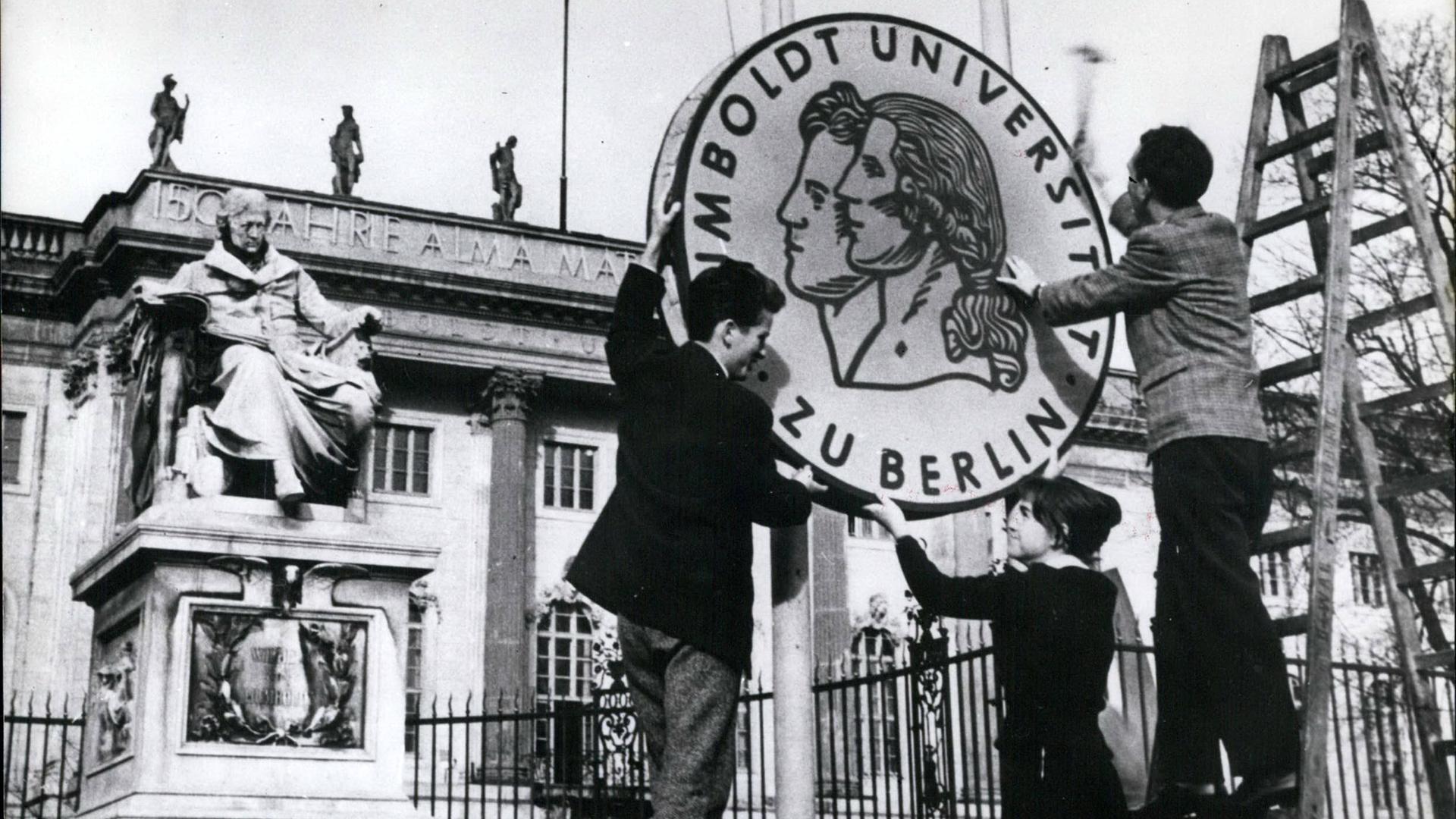 Drei Studierende halten vor der Humboldt-Uni zusammen ein großes, rundes Uni-Logo in der Hand.