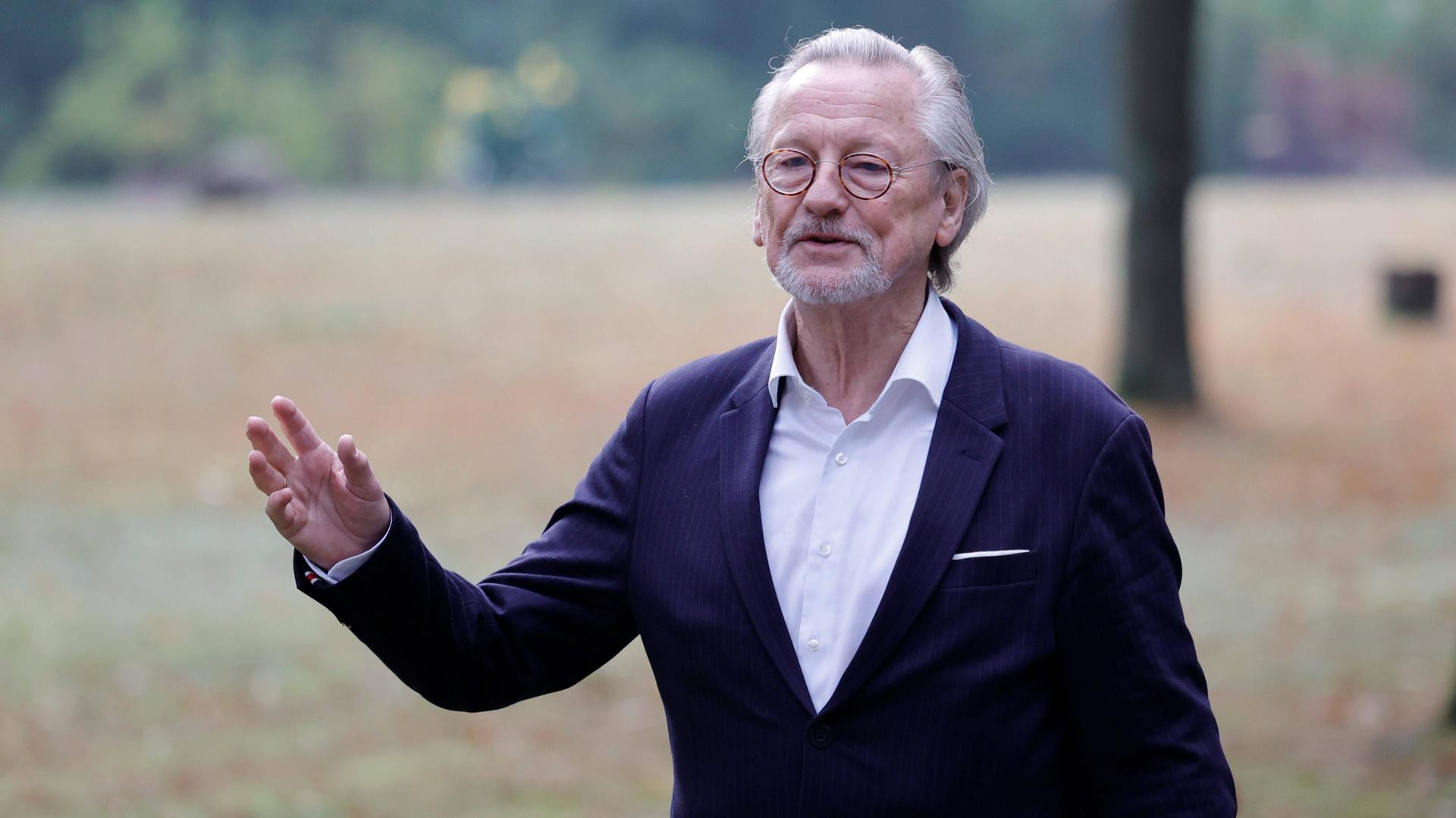 Peter Brandt trägt Anzug und ein weißes Hemd und steht gestikulierend auf einer Wiese. Peter Brandt trägt Anzug und ein weißes Hemd und steht gestikulierend auf einer Wiese.