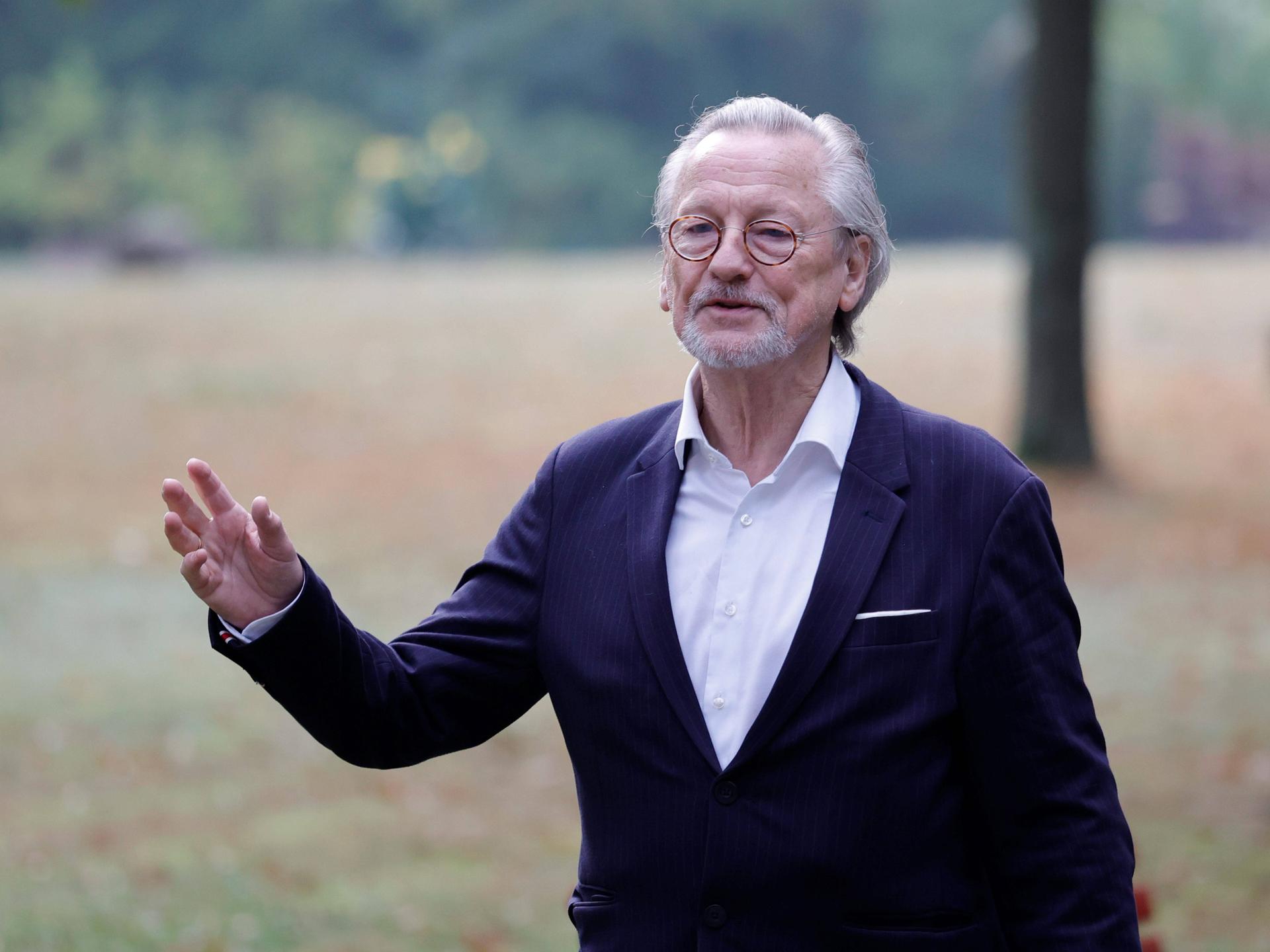 Peter Brandt trägt Anzug und ein weißes Hemd und steht gestikulierend auf einer Wiese.