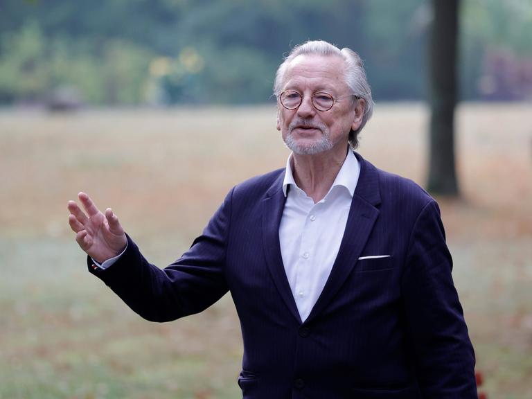 Peter Brandt trägt Anzug und ein weißes Hemd und steht gestikulierend auf einer Wiese.