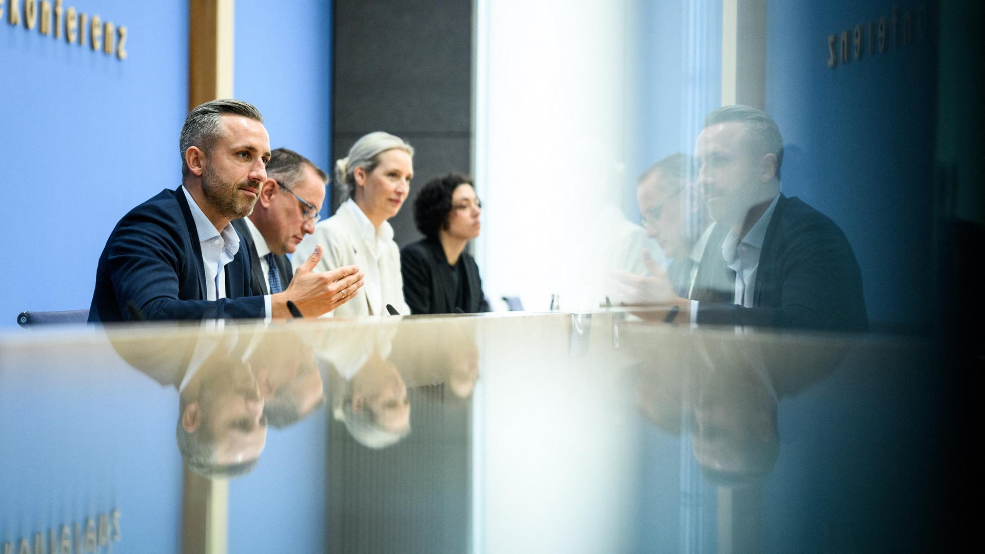 Ulrich Siegmund, Tino Chrupalla und Alice Weidel sitzen bei der Bundespressekonferenz an einem reflektierenden Tisch und vor einer Glastür. Auf beiden Oberflächen spiegeln sich die drei Personen. Ulrich Siegmund, Tino Chrupalla und Alice Weidel sitzen bei der Bundespressekonferenz an einem reflektierenden Tisch und vor einer Glastür. Auf beiden Oberflächen spiegeln sich die drei Personen.