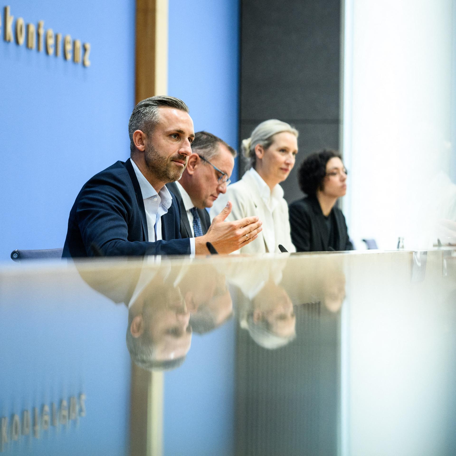 Ulrich Siegmund, Tino Chrupalla und Alice Weidel sitzen bei der Bundespressekonferenz an einem reflektierenden Tisch und vor einer Glastür. Auf beiden Oberflächen spiegeln sich die drei Personen.
