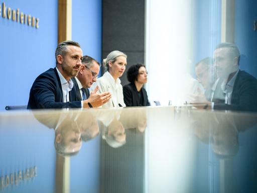 Ulrich Siegmund, Tino Chrupalla und Alice Weidel sitzen bei der Bundespressekonferenz an einem reflektierenden Tisch und vor einer Glastür. Auf beiden Oberflächen spiegeln sich die drei Personen.