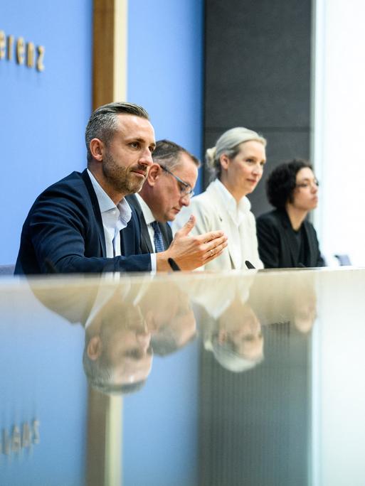 Ulrich Siegmund, Tino Chrupalla und Alice Weidel sitzen bei der Bundespressekonferenz an einem reflektierenden Tisch und vor einer Glastür. Auf beiden Oberflächen spiegeln sich die drei Personen.