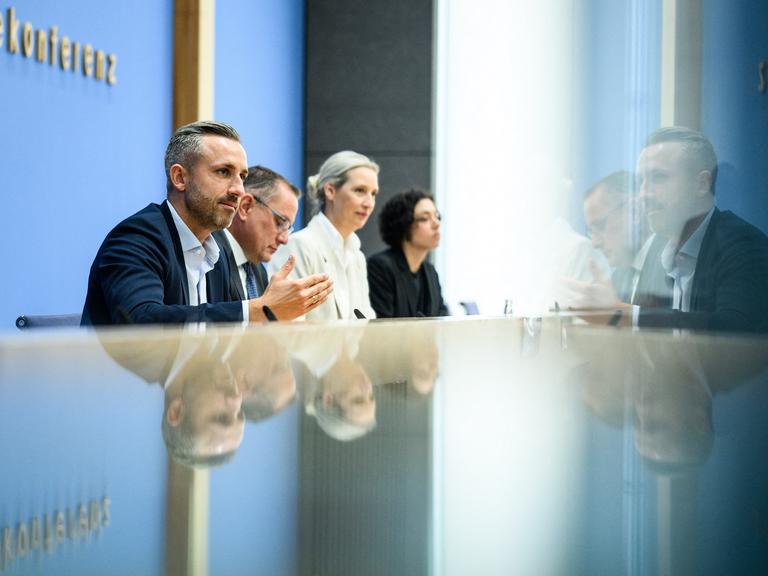 Ulrich Siegmund, Tino Chrupalla und Alice Weidel sitzen bei der Bundespressekonferenz an einem reflektierenden Tisch und vor einer Glastür. Auf beiden Oberflächen spiegeln sich die drei Personen. Ulrich Siegmund, Tino Chrupalla und Alice Weidel sitzen bei der Bundespressekonferenz an einem reflektierenden Tisch und vor einer Glastür. Auf beiden Oberflächen spiegeln sich die drei Personen.