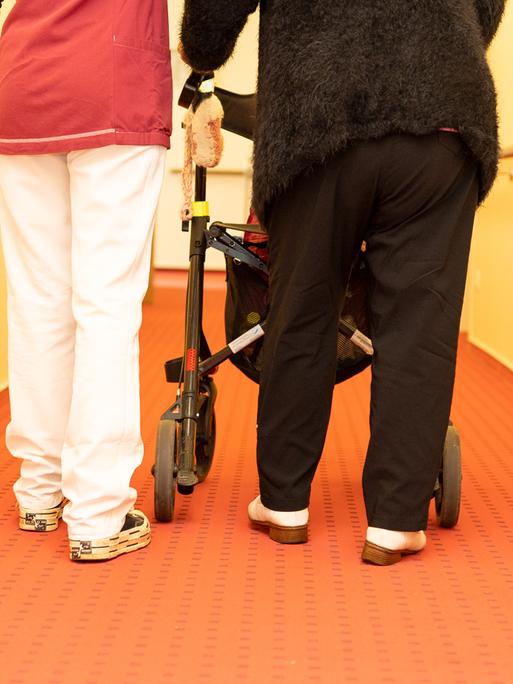 In einem Altenheim begleitet eine Pflegekraft eine Bewohnerin des Seniorenheims mit derem Rollator auf dem Flur der Einrichtung.