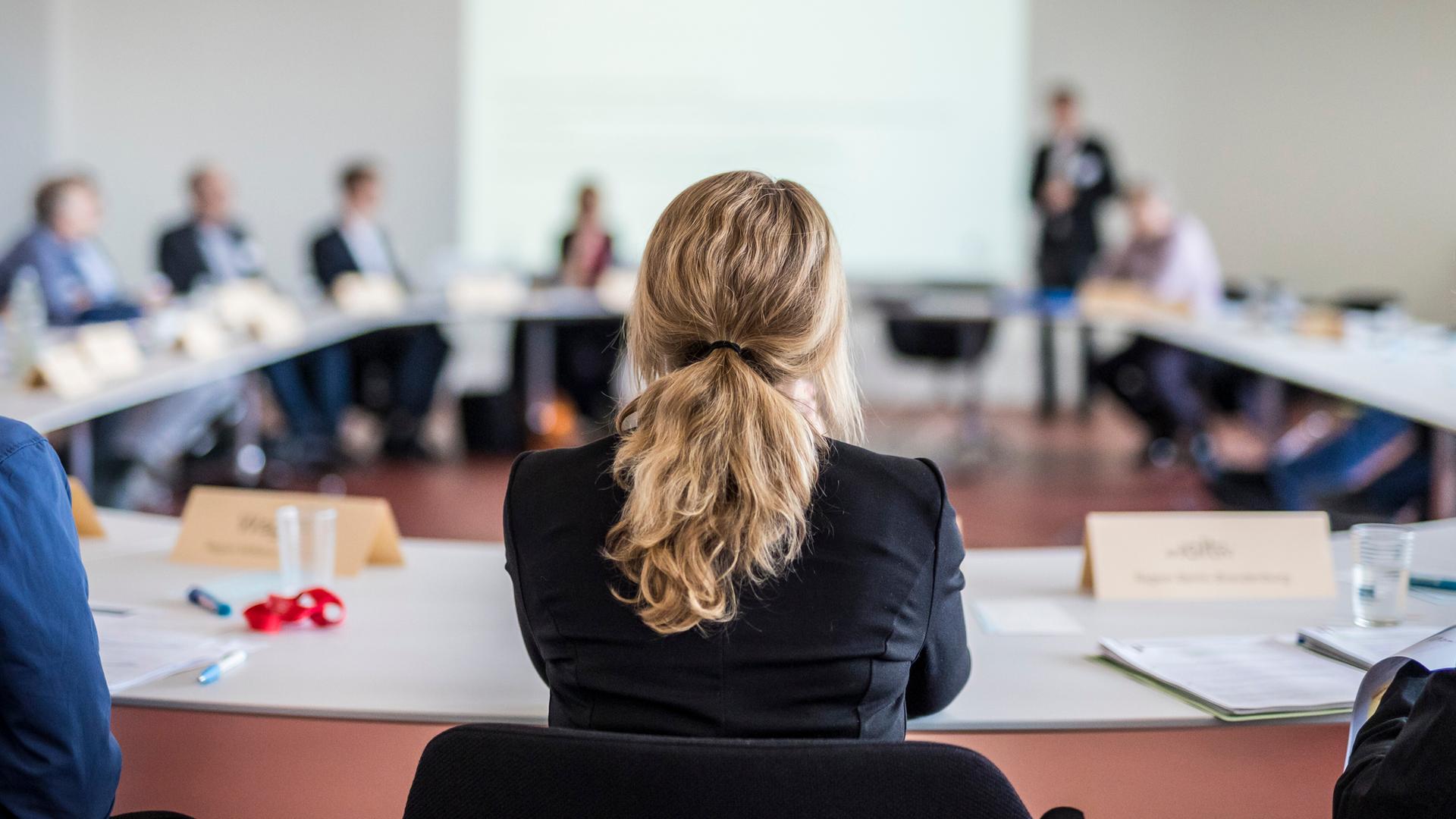Ein ovaler Arbeitstisch mit mehreren Personen. Direkt vorne sitzt eine Frau mit langen Haaren. Auf der anderen Seite steht eine Person und scheint etwas zu erklären. 