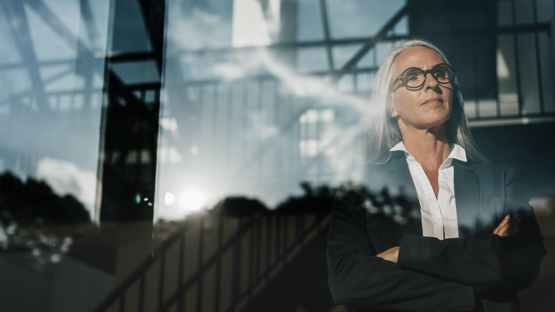 Eine Frau im schwarzen Blazer steht mit verschränkten Armen an einem Fenster und schaut mit entschlossenem Blick hinaus.