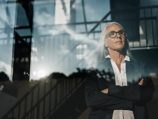 Eine Frau im schwarzen Blazer steht mit verschränkten Armen an einem Fenster und schaut mit entschlossenem Blick hinaus.