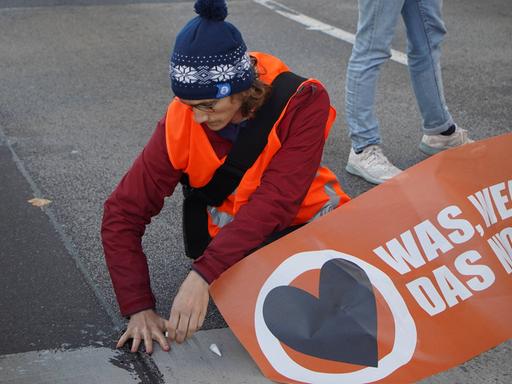 Ein Aktivist mit orangener Weste klebt sich am Asphalt fest.