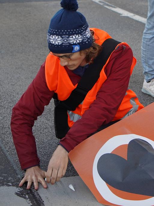 Ein Aktivist mit orangener Weste klebt sich am Asphalt fest.