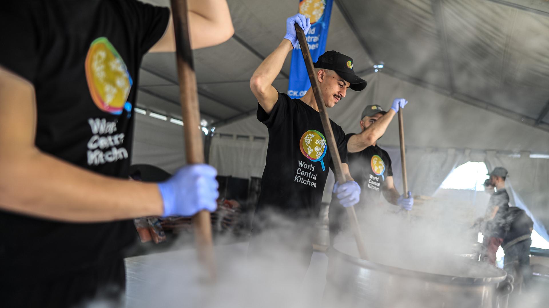 Drei Männer in schwarzen T-Shirts mit dem Aufdruck der Organisation rühren in einem Zelt mit langen Stangen in großen Kochtöpfen Essen um. Aus den Töpfen steigt dichter weißer Dampf auf.