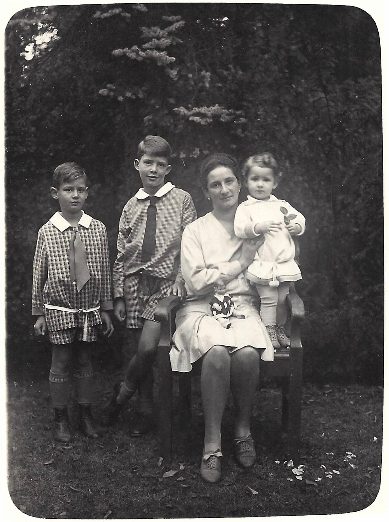 Schwarzweiß-Foto: Eine Frau im hellen Kleid sitzt draußen auf einem Stuhl und hält ihre kleine Tochter fest, daneben stehen zwei Jungen im Schulalter