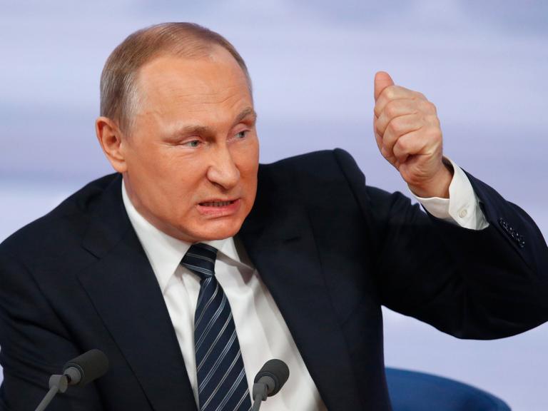 Porträt von Wladimir Putin vor zwei Mikrofonen. Er guckt böse, während er spricht und hat die Faust erhoben, als wolle er damit auf den Tisch hauen.