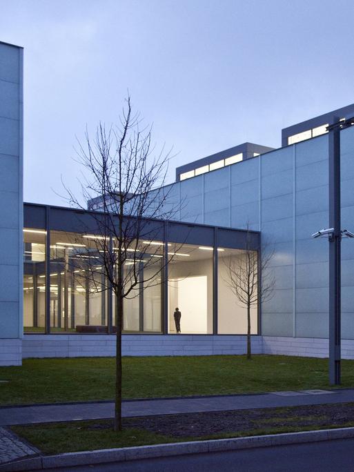 Außenaufnahme des Museums Folkwang in Essen vor Abendhimmel, im beleuchteten Gang, der zwei Gebäudeteile verbindet, ist die Silhouette eines Mannes zu sehen.