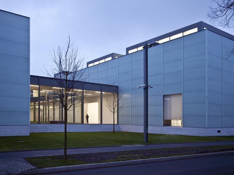 Außenaufnahme des Museums Folkwang in Essen vor Abendhimmel, im beleuchteten Gang, der zwei Gebäudeteile verbindet, ist die Silhouette eines Mannes zu sehen.