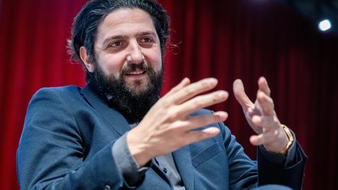 Aladin El-Mafaalani mit dunklen Haaren, Bart und im Anzug spricht und gestekuliert mit seinen Händen vor einem roten Vorhang