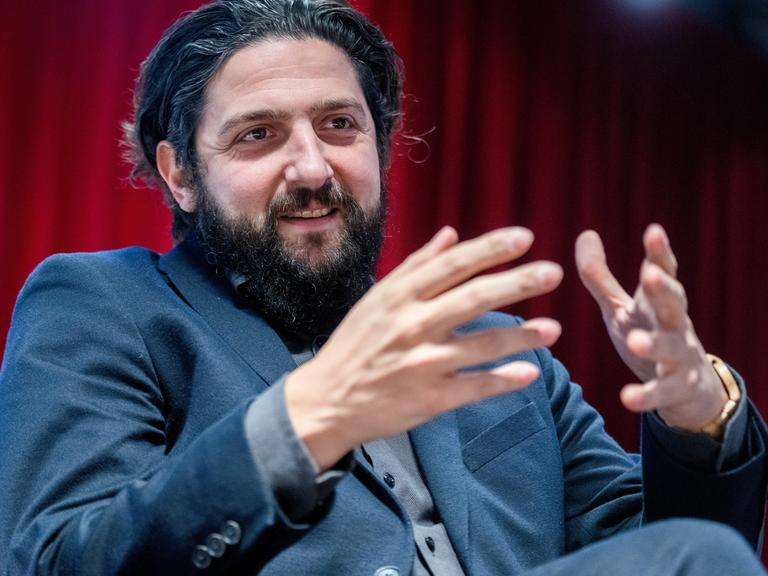 Aladin El-Mafaalani mit dunklen Haaren, Bart und im Anzug spricht und gestekuliert mit seinen Händen vor einem roten Vorhang