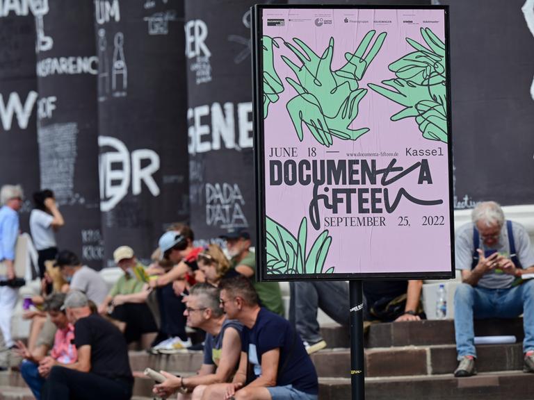 Besucher der documenta fifteen sitzen auf den Stufen des Fridericianums vor bemalten Säulen des Gebäudes; rechts ein Plakat der documenta
