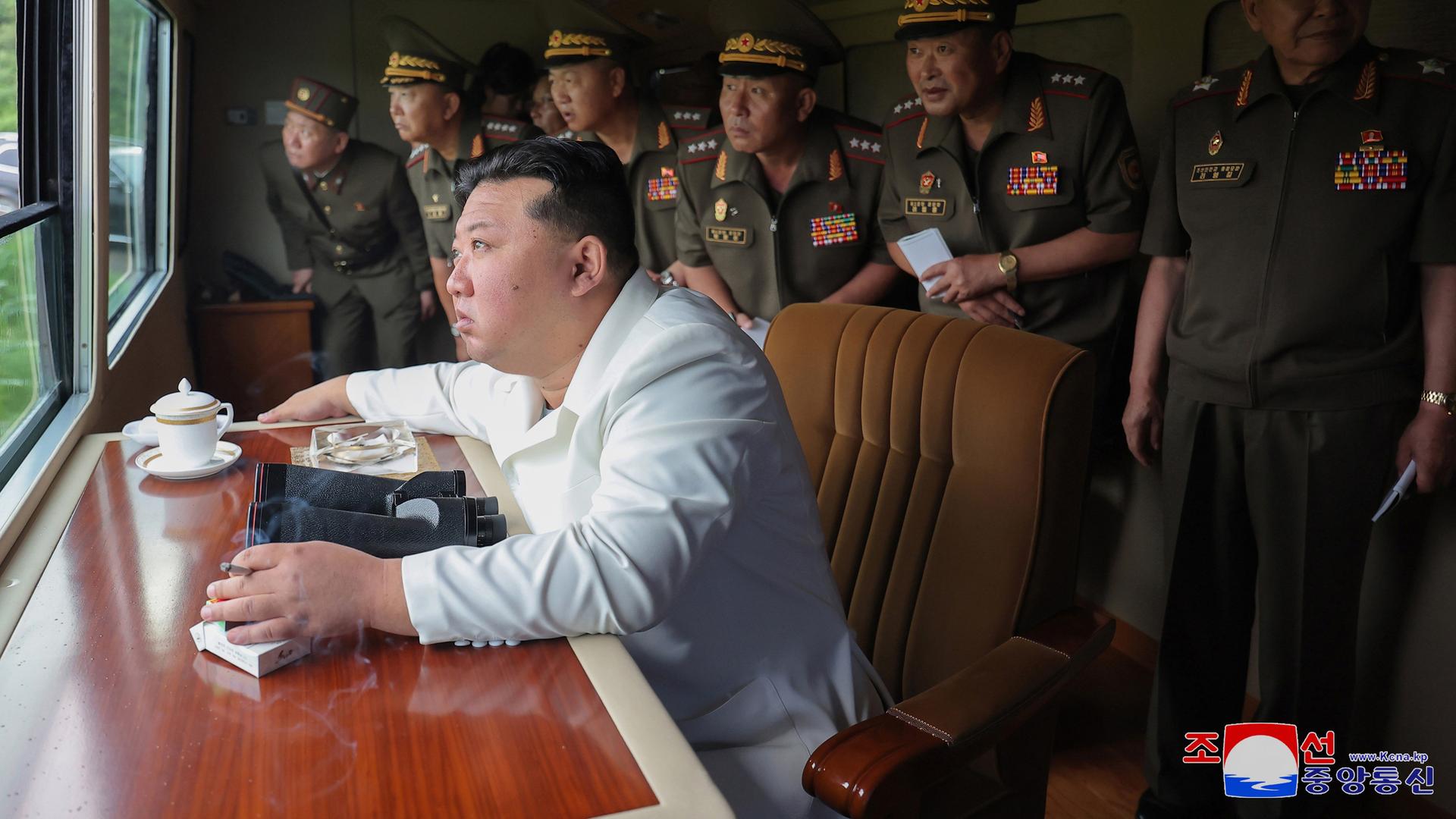 Nordkoreas Diktator Kim Jong-Un sitzt rauchend an einem Tisch und schaut aus einem Fenster, um den den Test eines neuen 240-Millimeter-Mehrfachraketenwerfer zu beobachten.