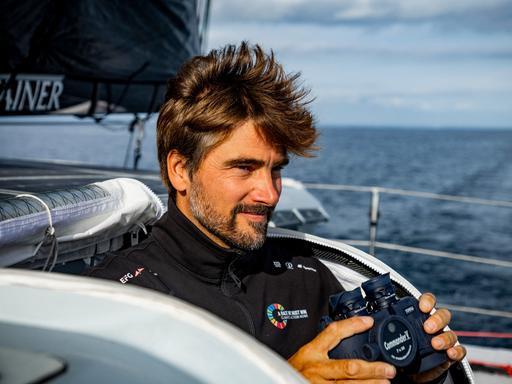 Boris Herrmann steht während einer Trainingsfahrt mit einem Fernglas in der Hand an Deck eines Segelbootes. Er ist ein mittelalter Mann mit braunem Haar und gestutztem Haar.