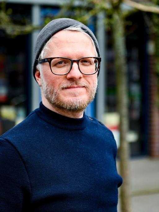 Verlagsleiter Daniel Beskos, Mitveranstalter des Indiebookday, einem Aktionstag zur Unterstützung kleiner unabhängiger Verlage, steht im Hamburger Schanzenviertel vor einer Buchhandlung. Er trägt eine Brille mit dunklem Gestell, Wollmütze und Fünf-Tage-Bart. 