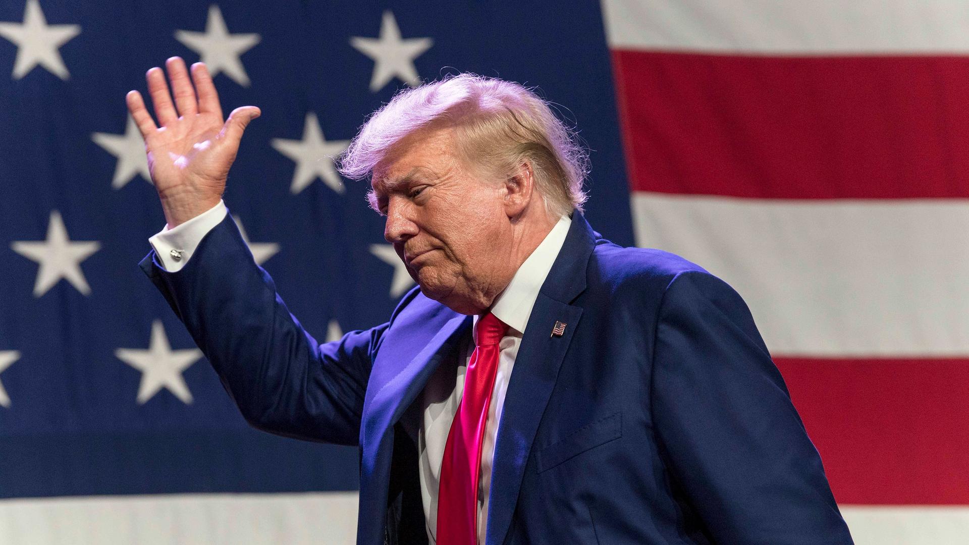 Donald Trump vor einer US-amerikanischen Flagge winkend im Abgang von einer Bühne. 