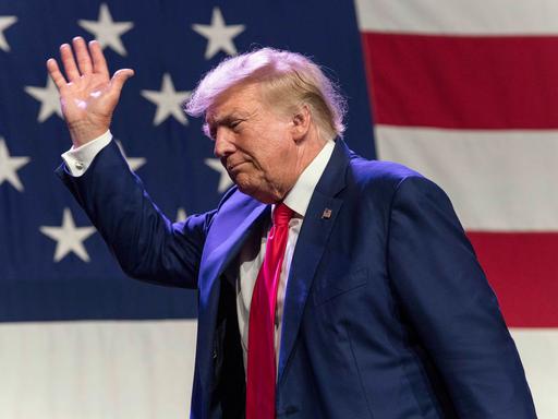 Donald Trump vor einer US-amerikanischen Flagge winkend im Abgang von einer Bühne. 