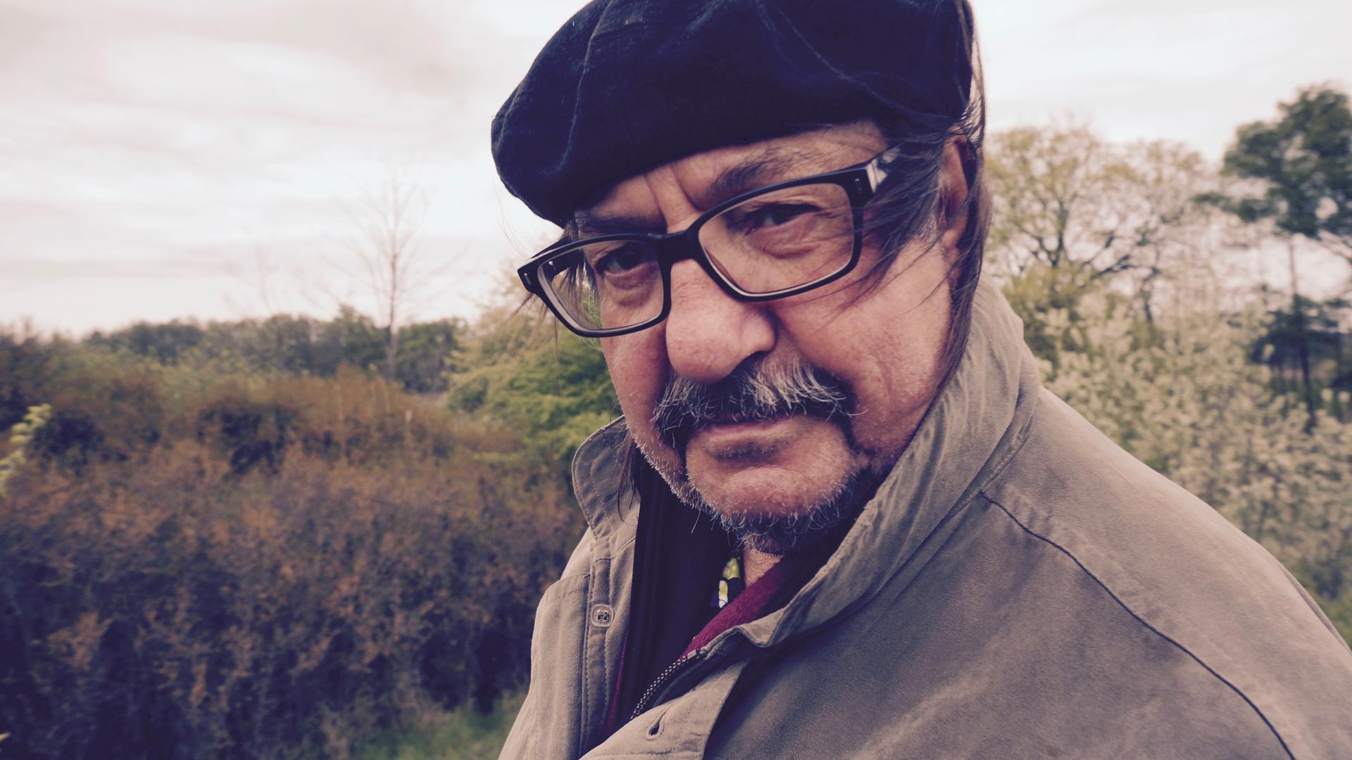 Der Autor und Liedermacher Manfred Maurenbrecher trägt Brille und Mütze und steht in der Natur. Der Wind bläst ihm die Haare ins Gesicht.