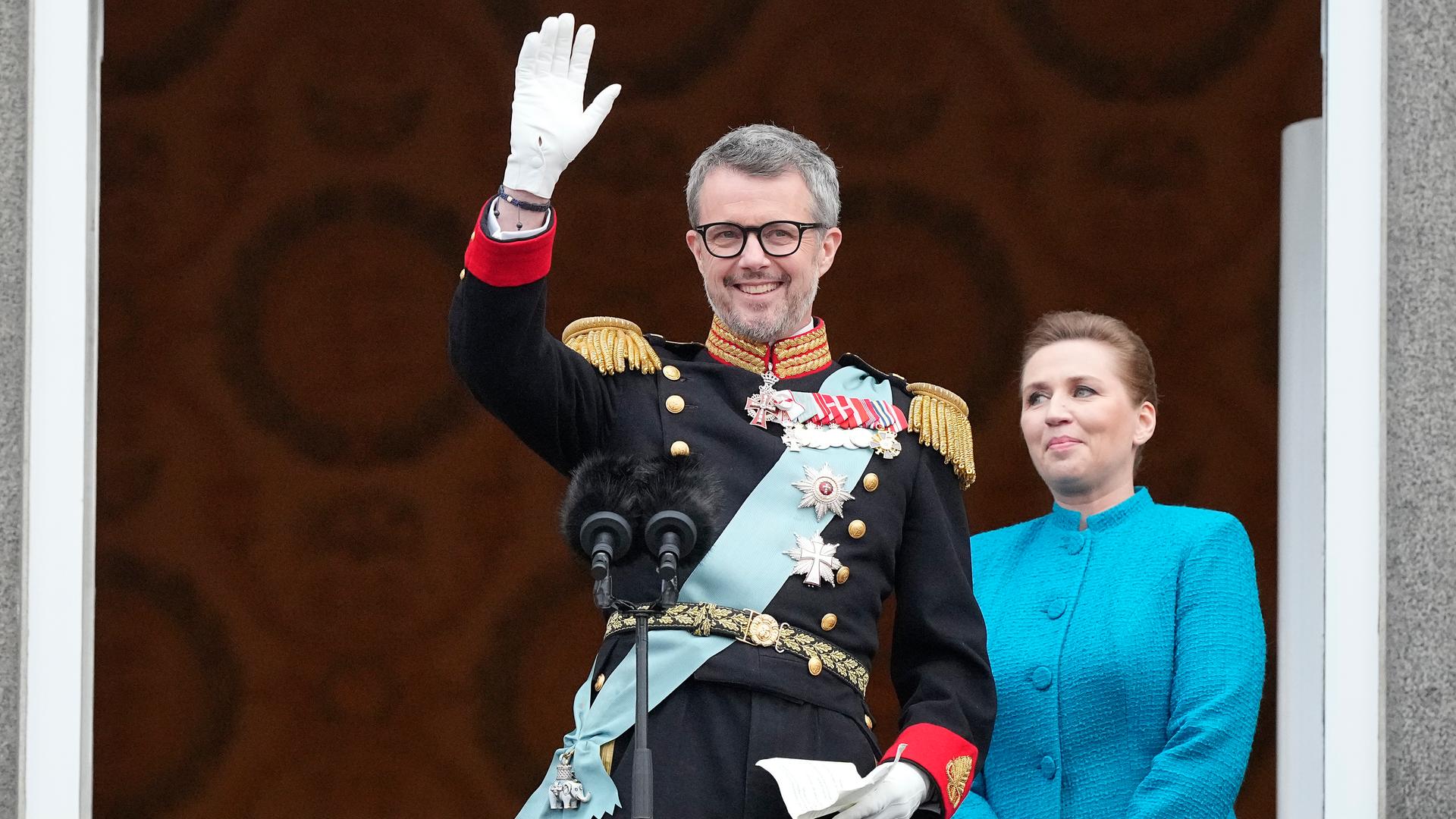 König Frederik X. steht auf dem Balkon von Schloss Christiansborg und winkt. Rechts hinter ihm steht die dänische Ministerpräsidentin Frederiksen.