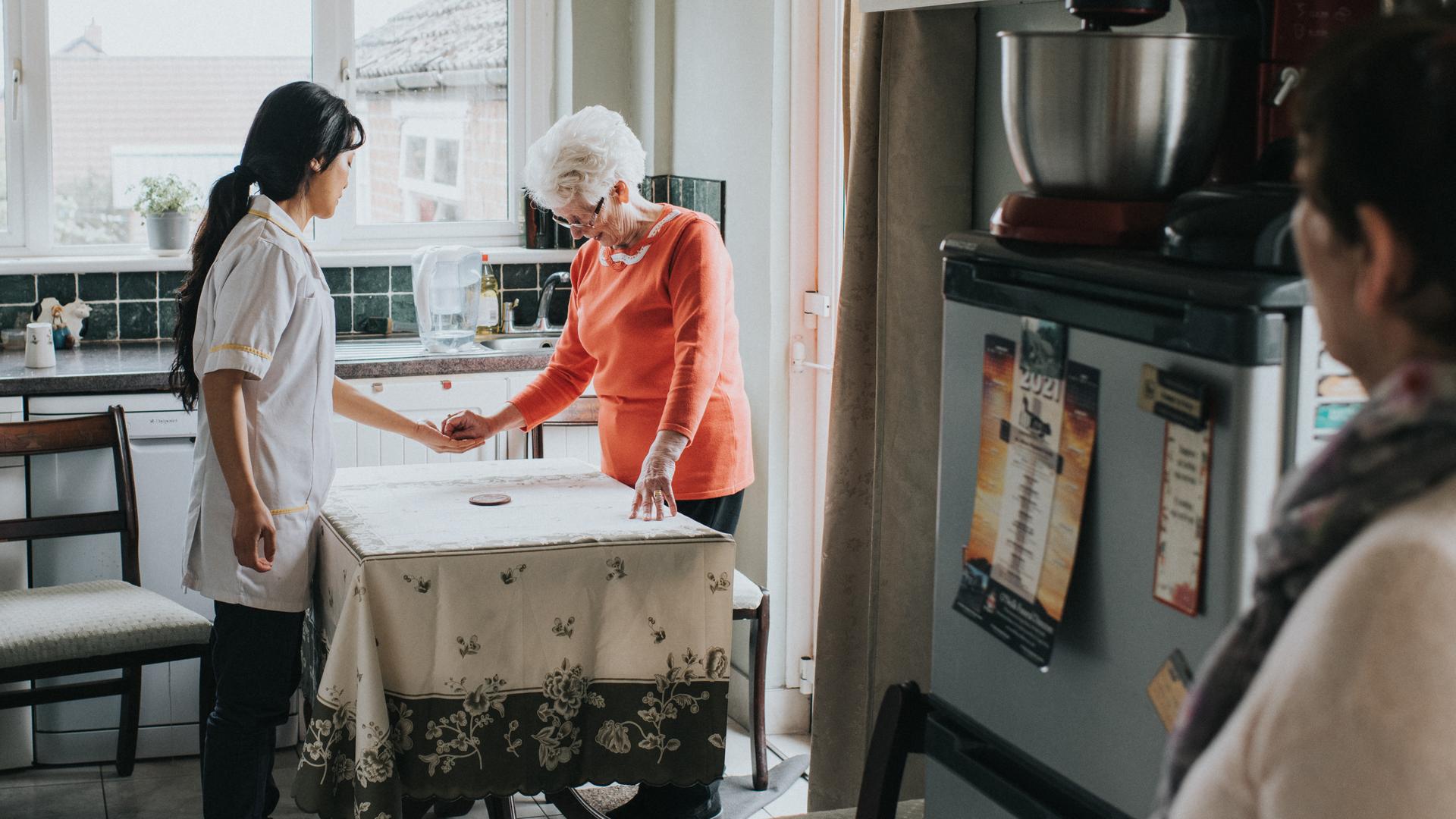 Eine ältere Dame steht mit ihrer Pflegekraft am Küchentisch und drückt ihr etwas in die Hand, während eine dritte Person mit etwas Abstand die Szene beobachtet.