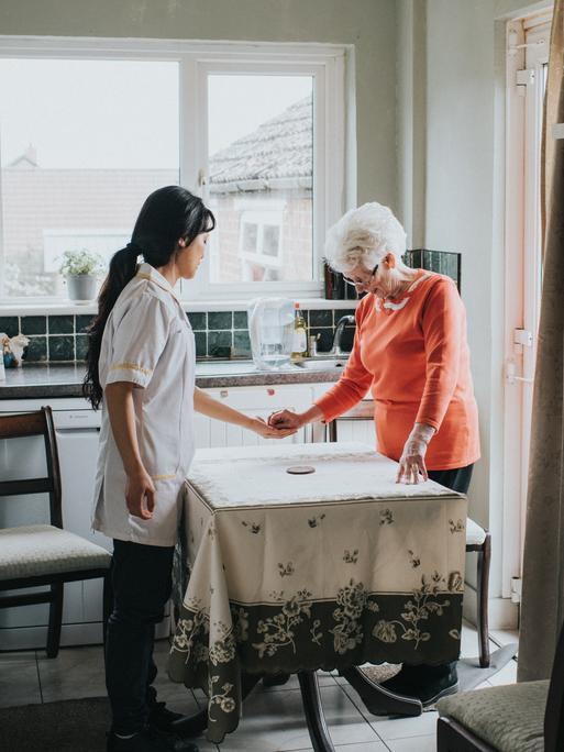Eine ältere Dame steht mit ihrer Pflegekraft am Küchentisch und drückt ihr etwas in die Hand, während eine dritte Person mit etwas Abstand die Szene beobachtet.