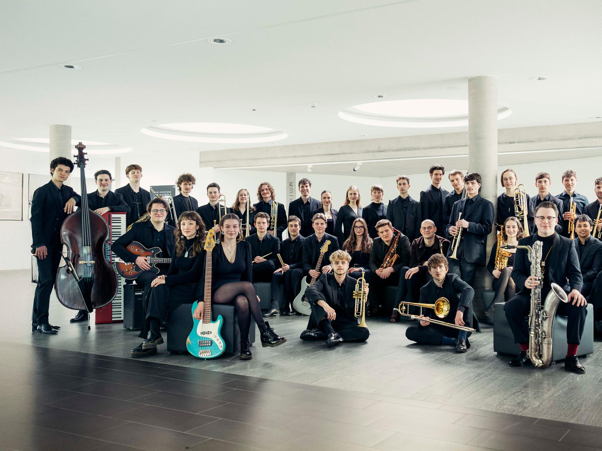 Die jungen Mitglieder des Orchesters stehen mit ihren Instrumenten in einem hellen, modernen Raum mit großen, runden Deckenleuchten.