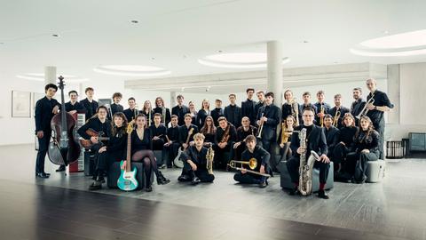 Die jungen Mitglieder des Orchesters stehen mit ihren Instrumenten in einem hellen, modernen Raum mit großen, runden Deckenleuchten.
