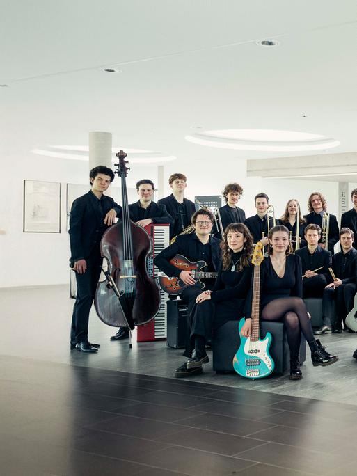 Die jungen Mitglieder des Orchesters stehen mit ihren Instrumenten in einem hellen, modernen Raum mit großen, runden Deckenleuchten.