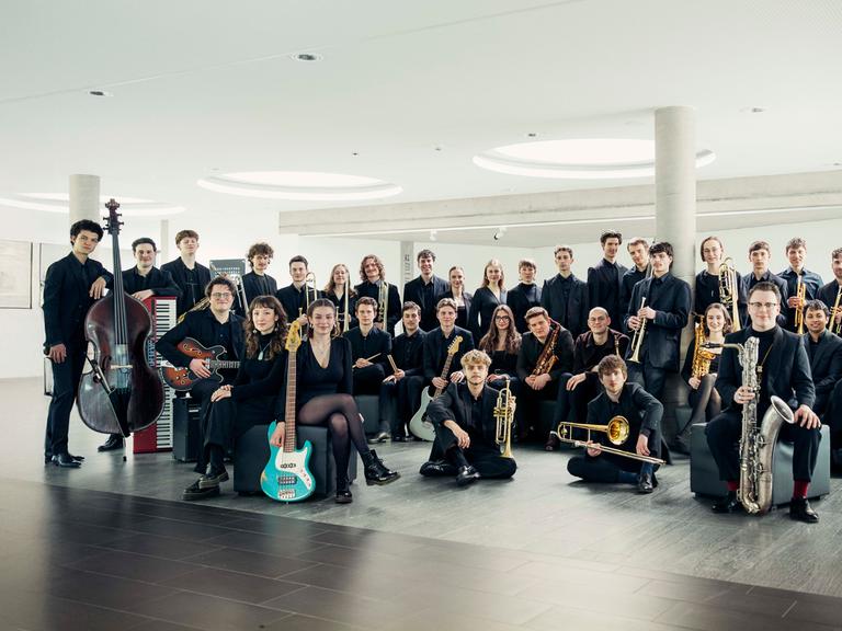 Die jungen Mitglieder des Orchesters stehen mit ihren Instrumenten in einem hellen, modernen Raum mit großen, runden Deckenleuchten.