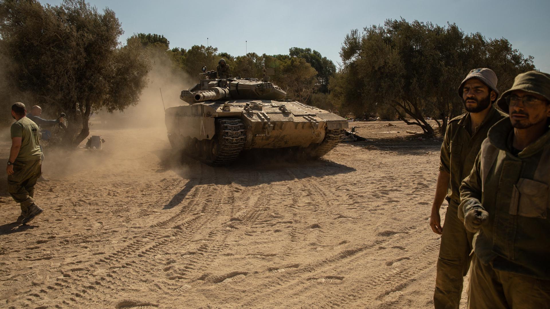 Ein israelischer Panzer und mehrere Soldaten auf staubigem Boden unter blauem Himmel.