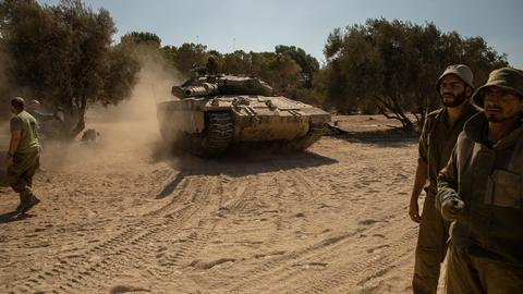 Ein israelischer Panzer und mehrere Soldaten auf staubigem Boden unter blauem Himmel.