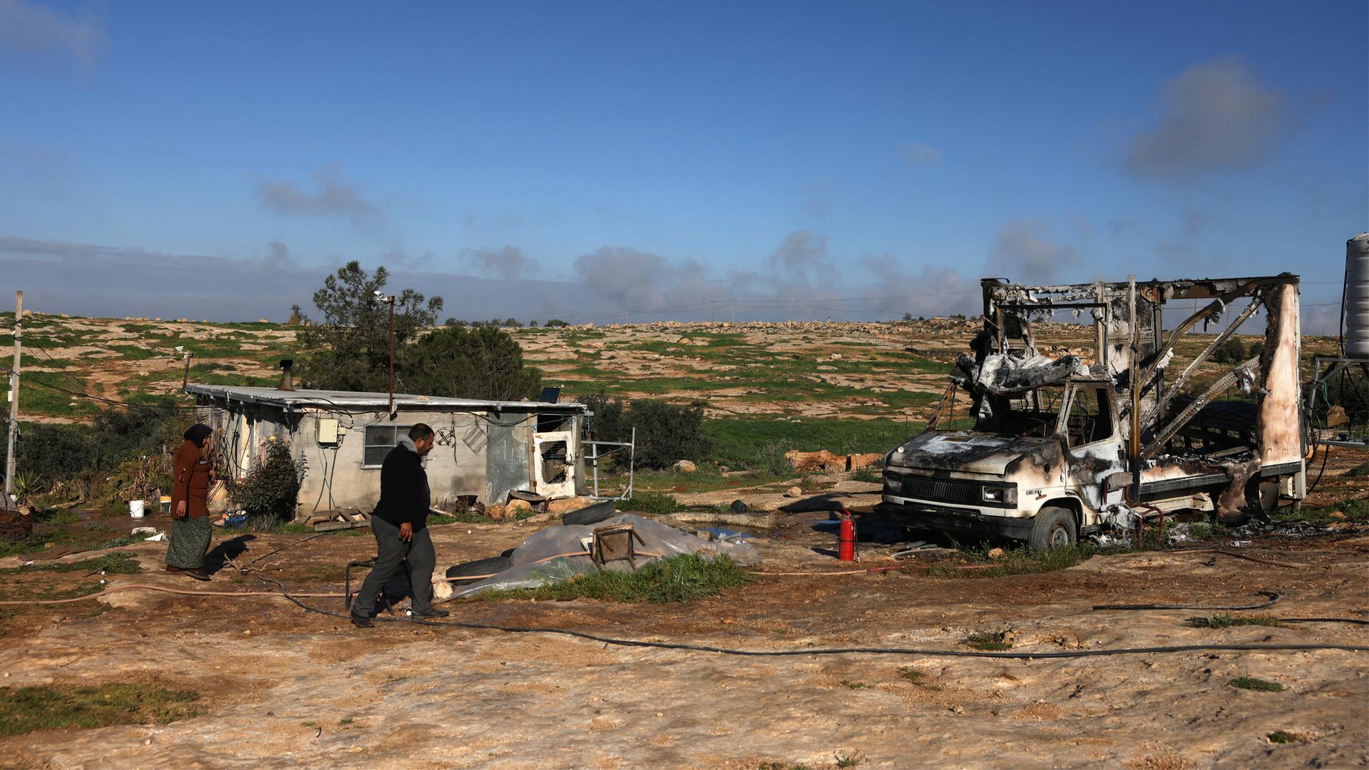 Ein Palästinenser geht an der ausgebrannten Karosserie eines Autos vorbei. Im Hintergrund ist staubiges Land zu sehen. 