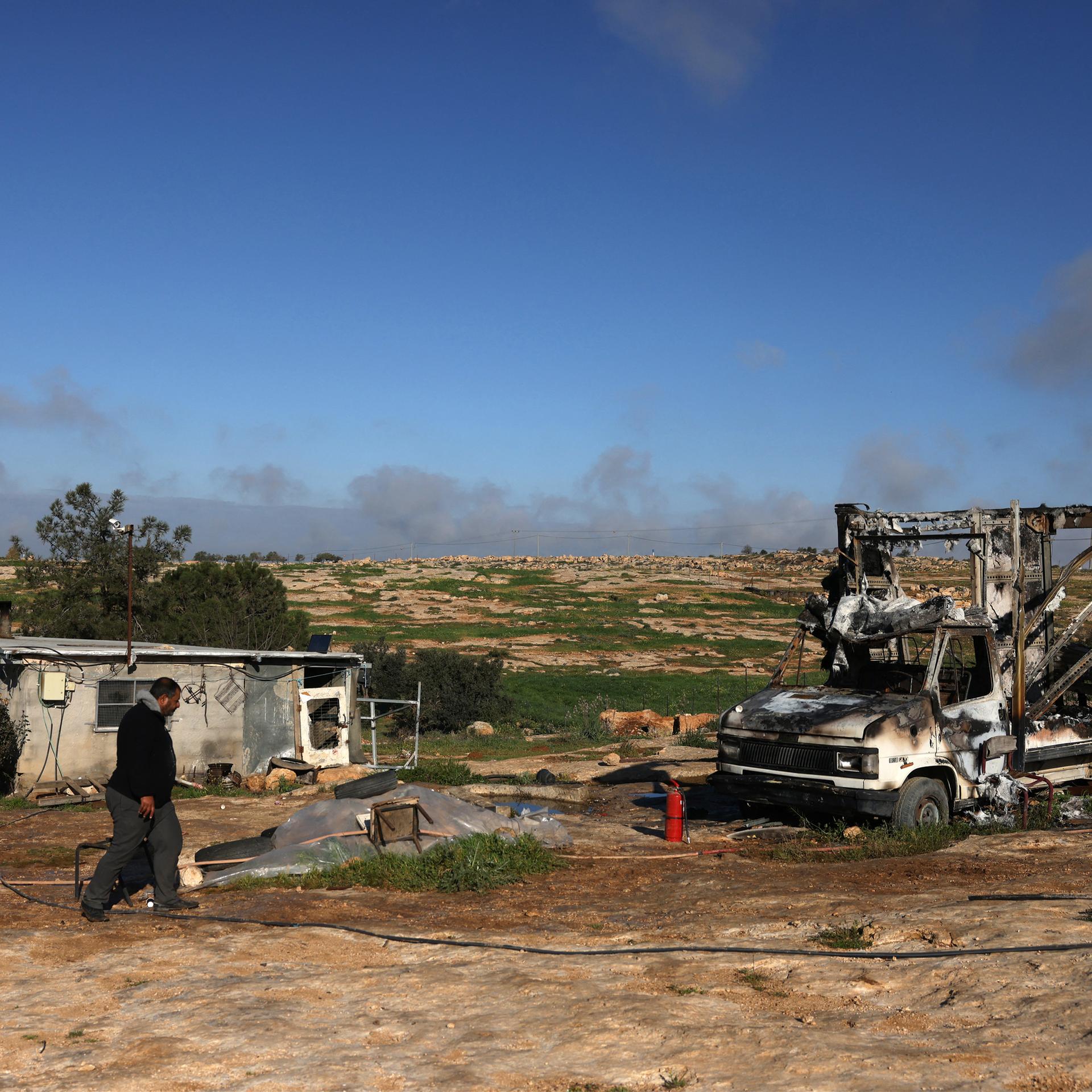 Ein Palästinenser geht an der ausgebrannten Karosserie eines Autos vorbei. Im Hintergrund ist staubiges Land zu sehen. 