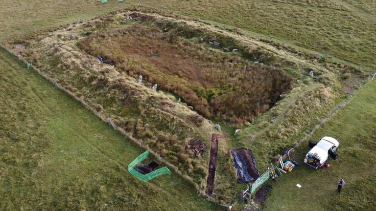 Luftaufnahme des Monuments "King Arthur's Hall": eine von Gras überwuchertes rechteckige Struktur mit Steinbegrenzungen.