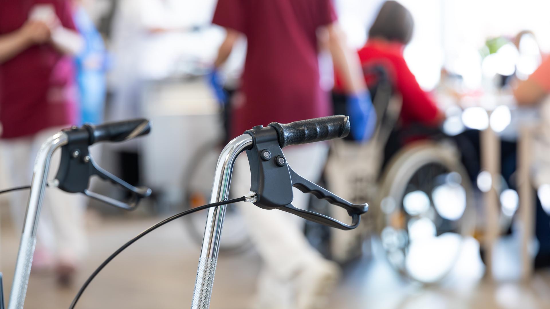 Ein Rollator steht in einem Seniorenheim, im Hintergrund sitzen ältere Menschen an einem Tisch.