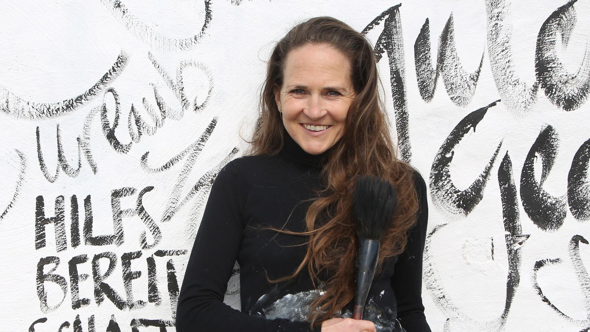 Die Künstlerin Jeannine Platz lehnt an einer weißen Wand, die sie mit Zeichen, Buchstaben und Wörtern bemalt hat. Sie trägt einen schwarzen Pulli und Jeans. 



