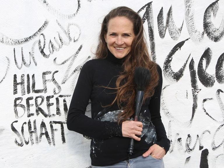 Die Künstlerin Jeannine Platz lehnt an einer weißen Wand, die sie mit Zeichen, Buchstaben und Wörtern bemalt hat. Sie trägt einen schwarzen Pulli und Jeans. 



