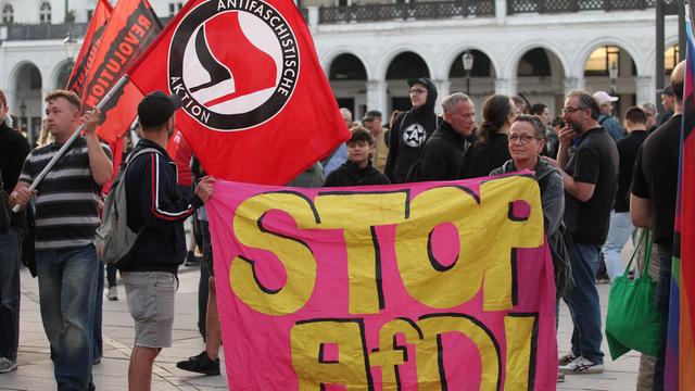 Zwei Menschen stehen auf einem Platz und halten während einer Demonstration einen Banner mit der Aufschrift " Stop AfD". Im Hintergrund befinden sich weitere Demonstrierende.