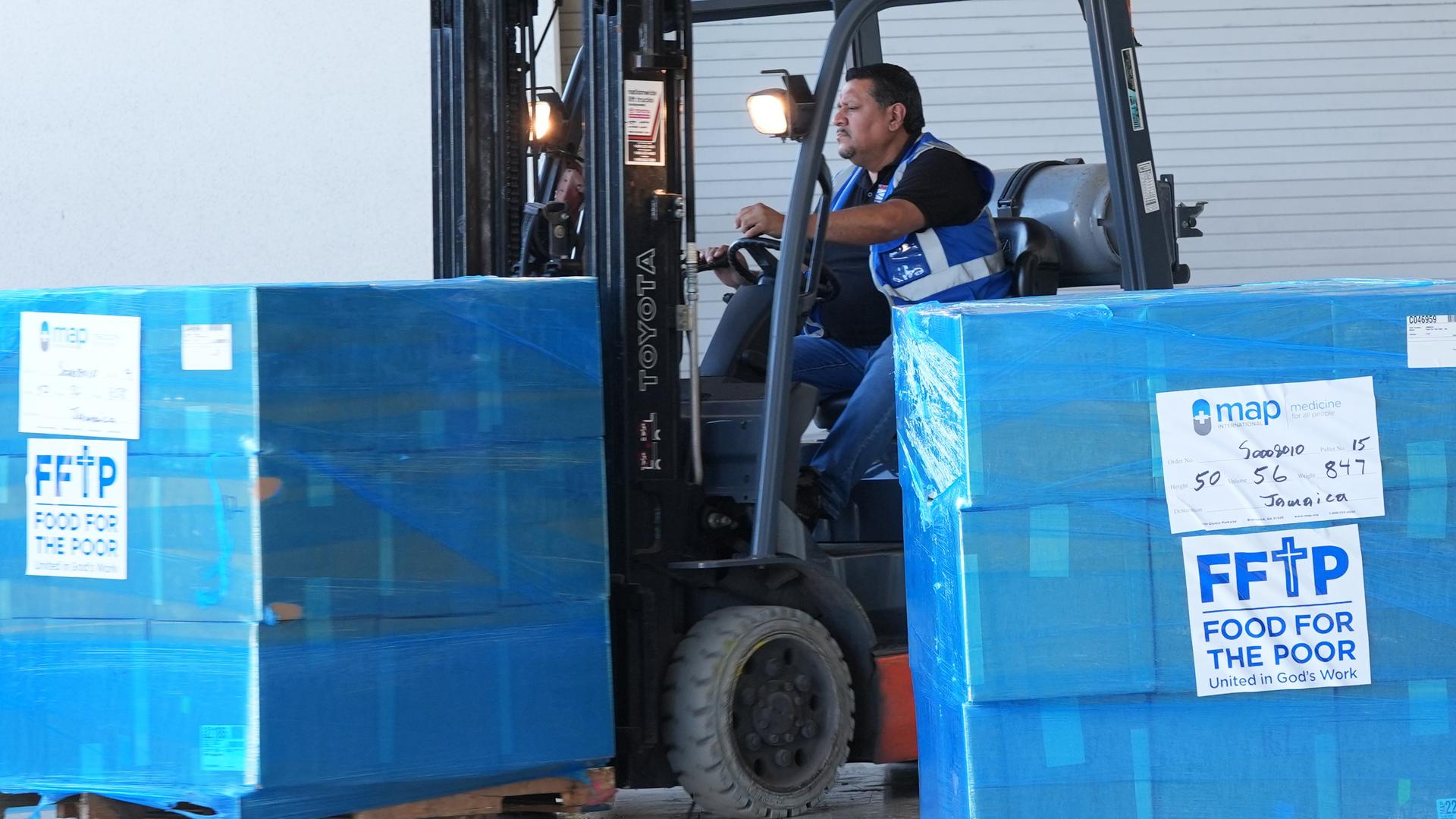 Ein Gabelstabler transportiert große blaue Kisten, auf denen steht "FFTP - Food for the poor". Ein Gabelstabler transportiert große blaue Kisten, auf denen steht "FFTP - Food for the poor".