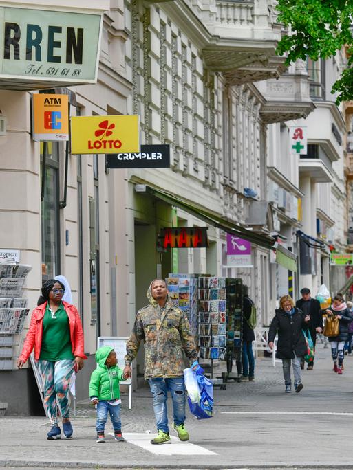 Eine Straßenszene in Berlin-Kreuzberg: Zwei dunkelhäutige Erwachsene und ein kleines Kind wollen eine Straße überqueren. Weitere Personen laufen auf dem Gehsteig.