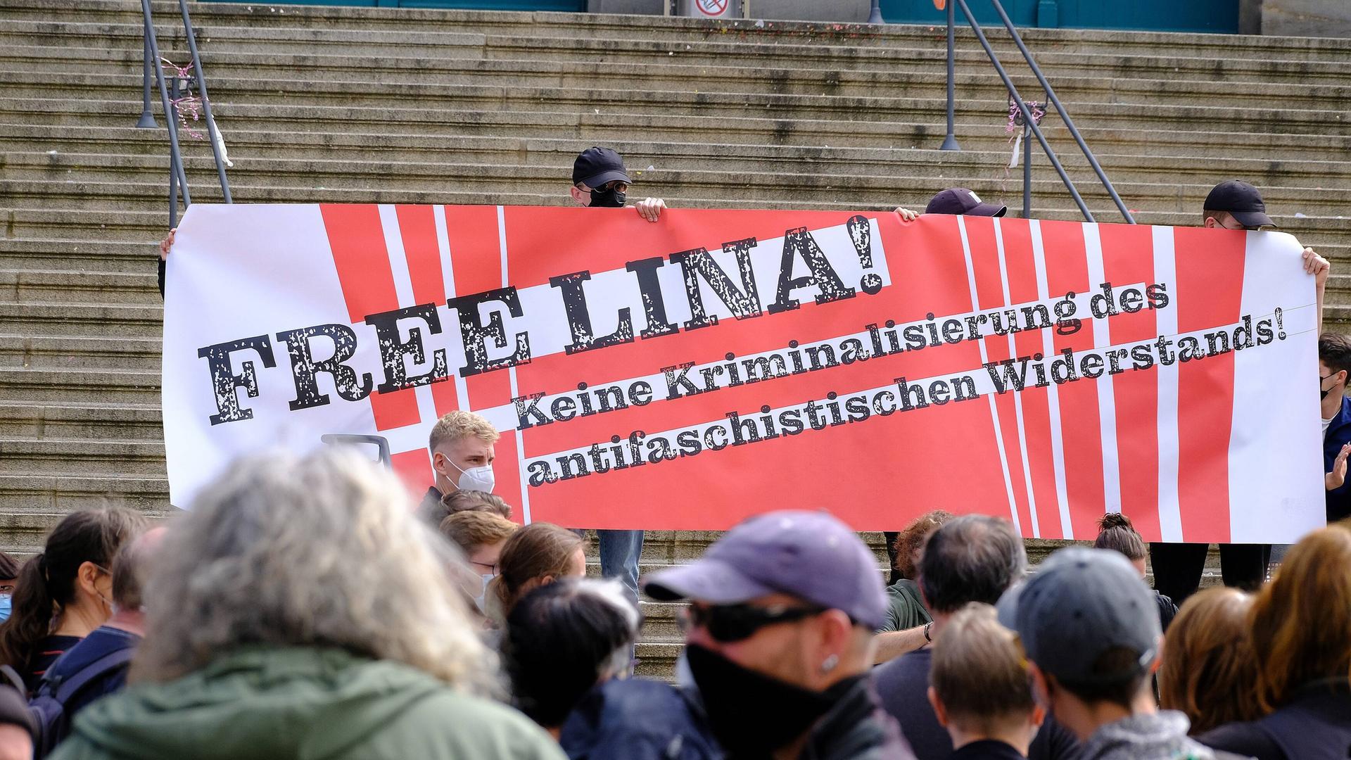 Menschen halten vor Demonstranten am Fuße einer Steintreppe ein großes Plakat hoch, auf dem steht "Free Lina! Keine Kriminalisierung des antifaschistischen Widerstands!"