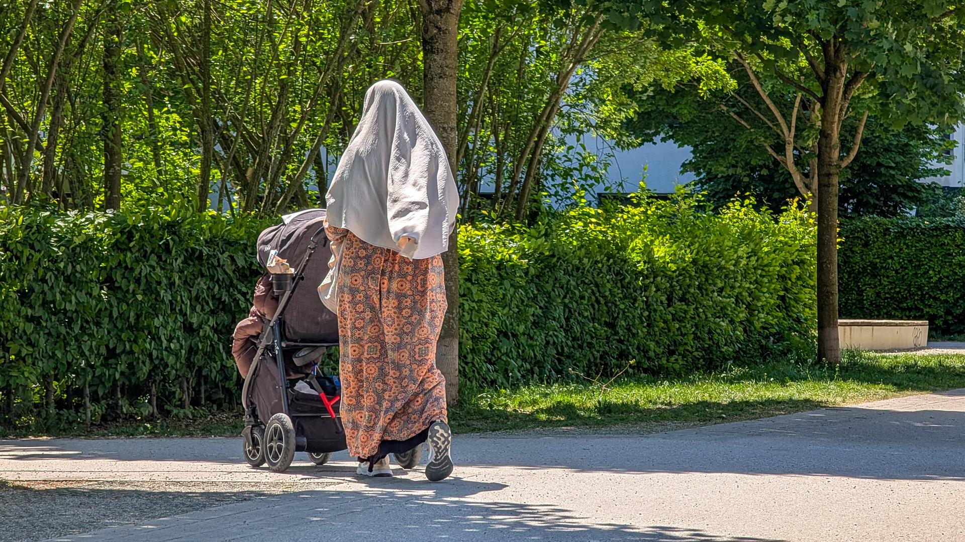 Eine Frau mit hellem Kopftuch und gemustertem Kleid schiebt einen Kinderwagen durch einen Park Eine Frau mit hellem Kopftuch und gemustertem Kleid schiebt einen Kinderwagen durch einen Park