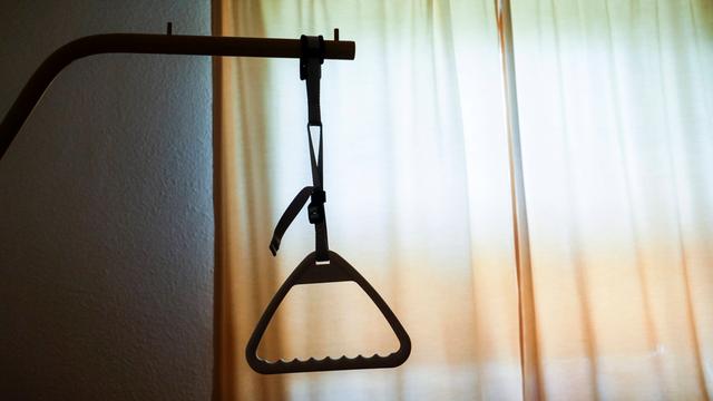 Der Armgriff eines Pflegebettes hebt sich als dunkle Silhouette vor einem Fenster ab.